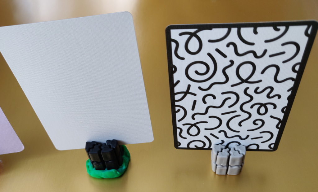 Two cards are shown inserted into the TI'TAINS card holder.  The first is a plain blank white playing inserted into a black TI'TAIN that is adhered to a surface using green play-doh.  The other is a black and white patterned playing card inserted into a grey TI'TAINS base.