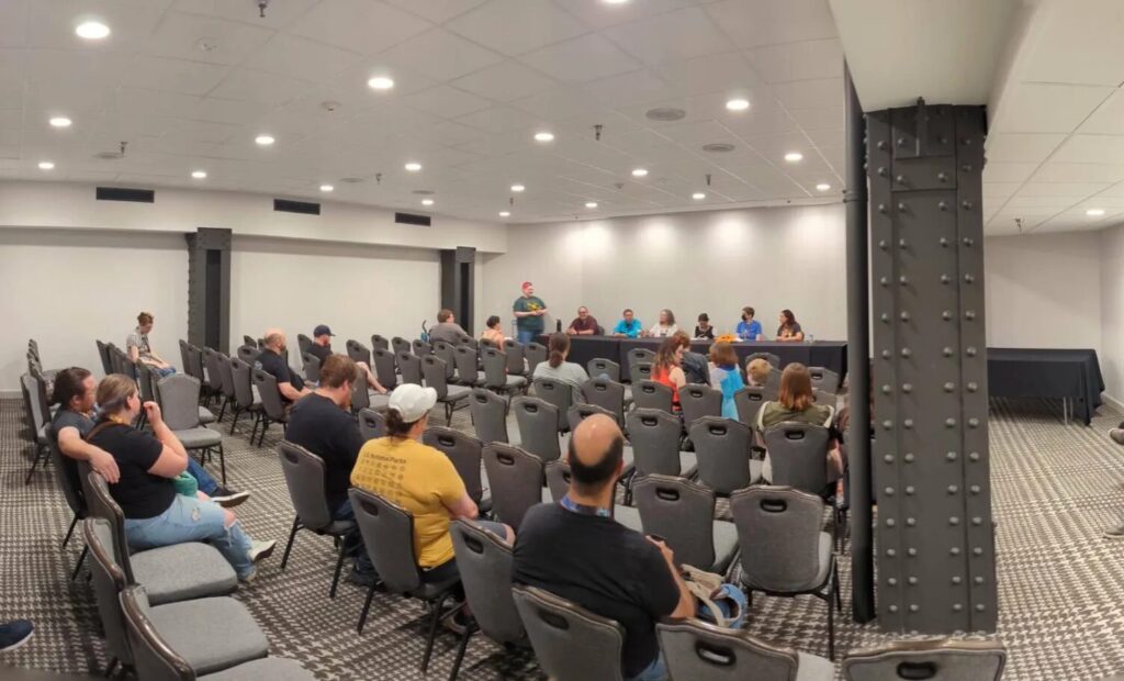 A photo of a conference room with folks sitting in rows of seats and 6 people sitting at a table at the far end of the room.  One person (Kat) is standing next to the table.