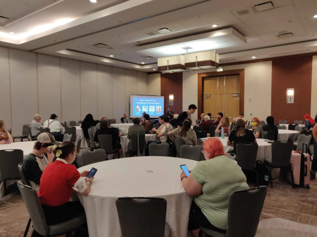 A conference room filled with round tables.  People are sitting at the tables, and there is a projector screen at the head of the room.