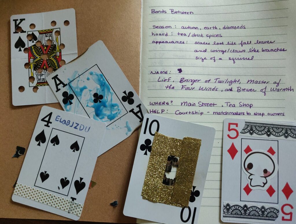 an image of an open notebook overlaid with 5 playing cards all modified in some way - one has holes punched in it, another has a hole burned in it's center and surrounded by gold tape, another is splattered with blue wax, another has a code written on it, and another has a ghost sticker at its center.
