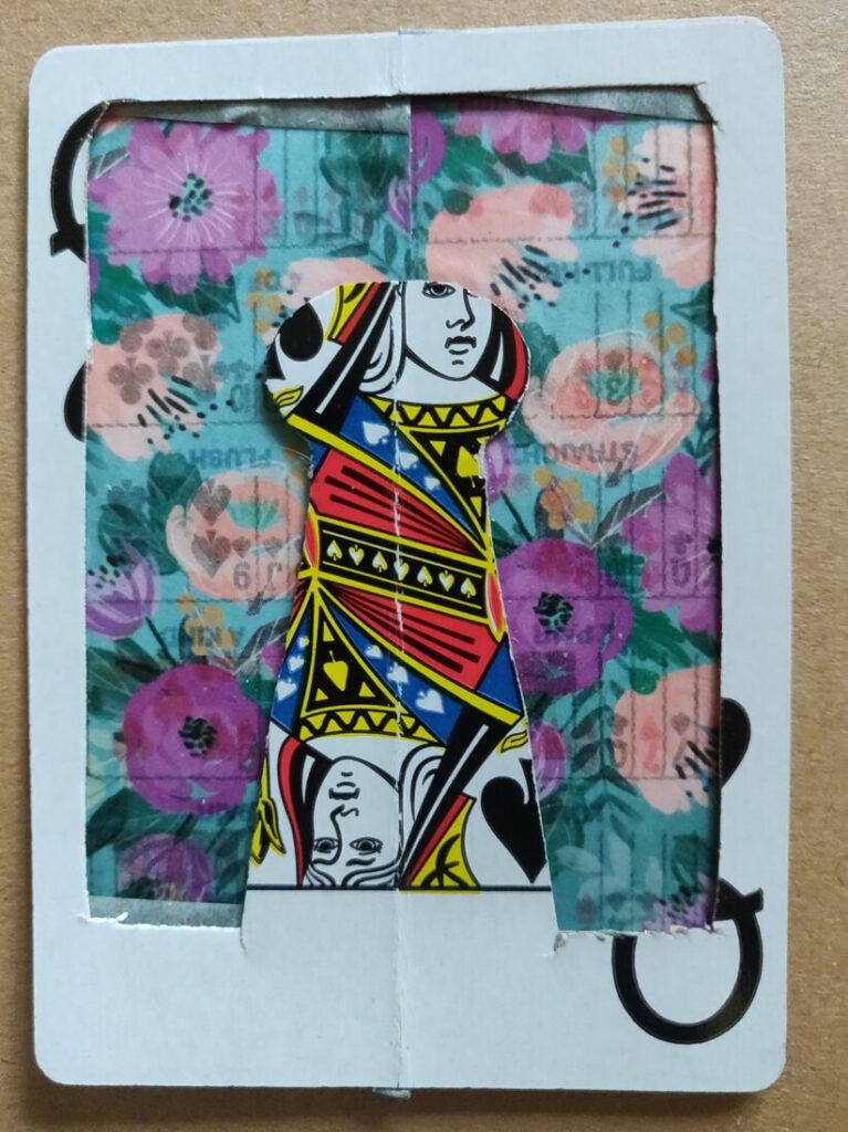 A Q of spades playing card that's cut to look like a game piece (like a pawn) in the center and has a flowery taped background behind it.