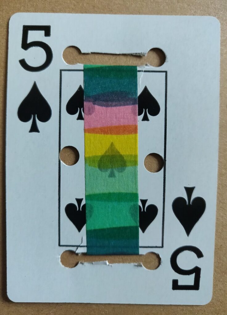 a 5 of spades playing card with holes punched in it and a piece of rainbow tape pasted over the center between two longer cuts in the card.