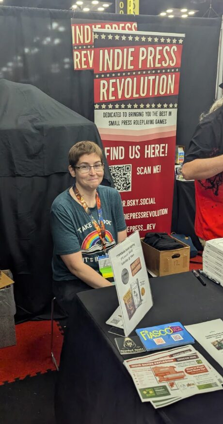 A pic of Steph from TTRPGkids sitting behind the IPR booth and smirking at noticing the camera guy