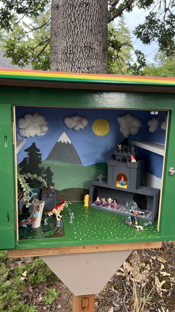 a photo of the interior of a little library box that contains a dioarama full of minis, a painted background scene, and a little castle