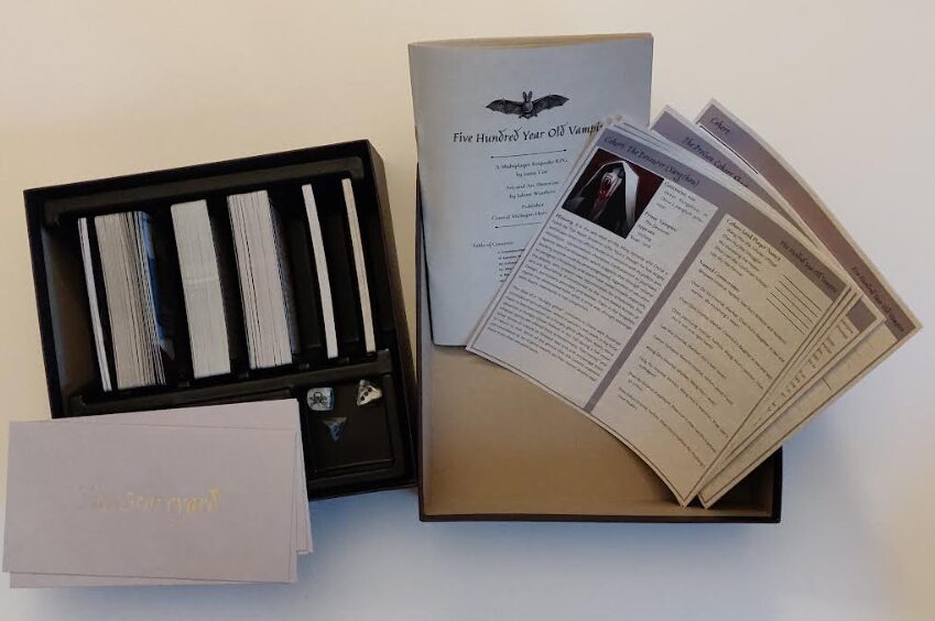 an open box for 500 Year Old Vampire displaying cards, dice, envelopes, pre-gen character cohort sheets, and a game book pamphlet