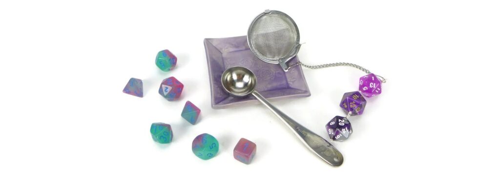 dice next to a tea infuster with three d20s linked on the infuser's chain. The infuser sits on a small dish along with a teaspoon.