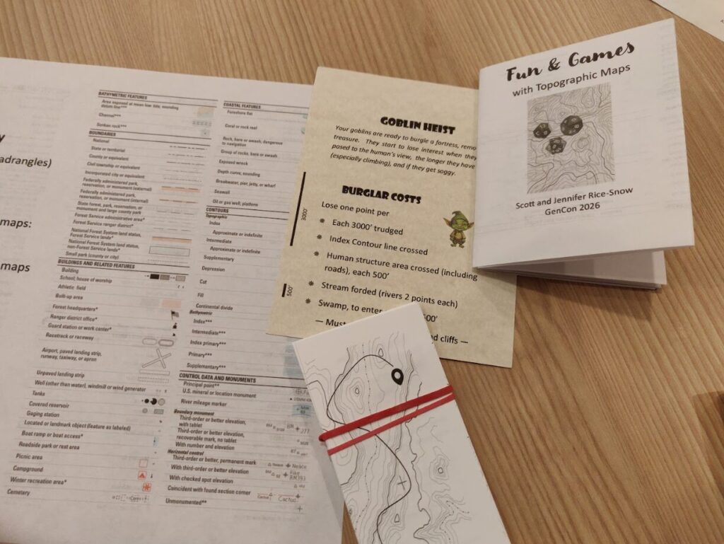 a photo of handouts and game materials for the fun and games with topographic maps talk. 

These materials include a set of cards, a zine called "fun and games with topographic maps by Scott and Jennifer Rice-Snow" and the Goblin Heist game card